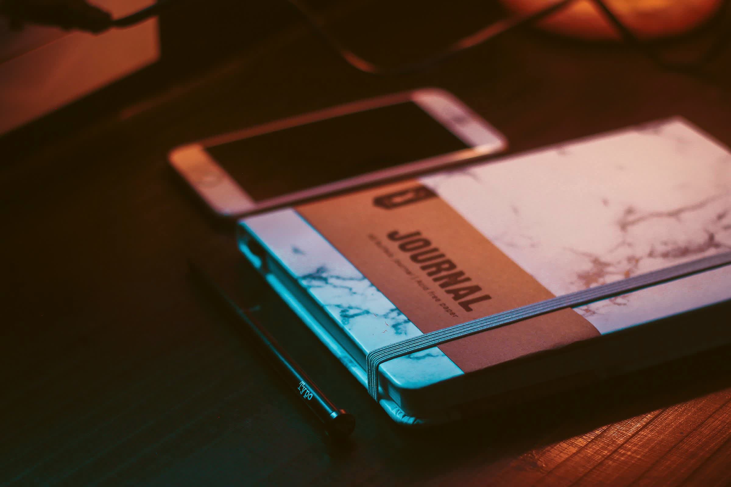 A paper journal, a phone and a pen resting on a dark wooden table by candlelight